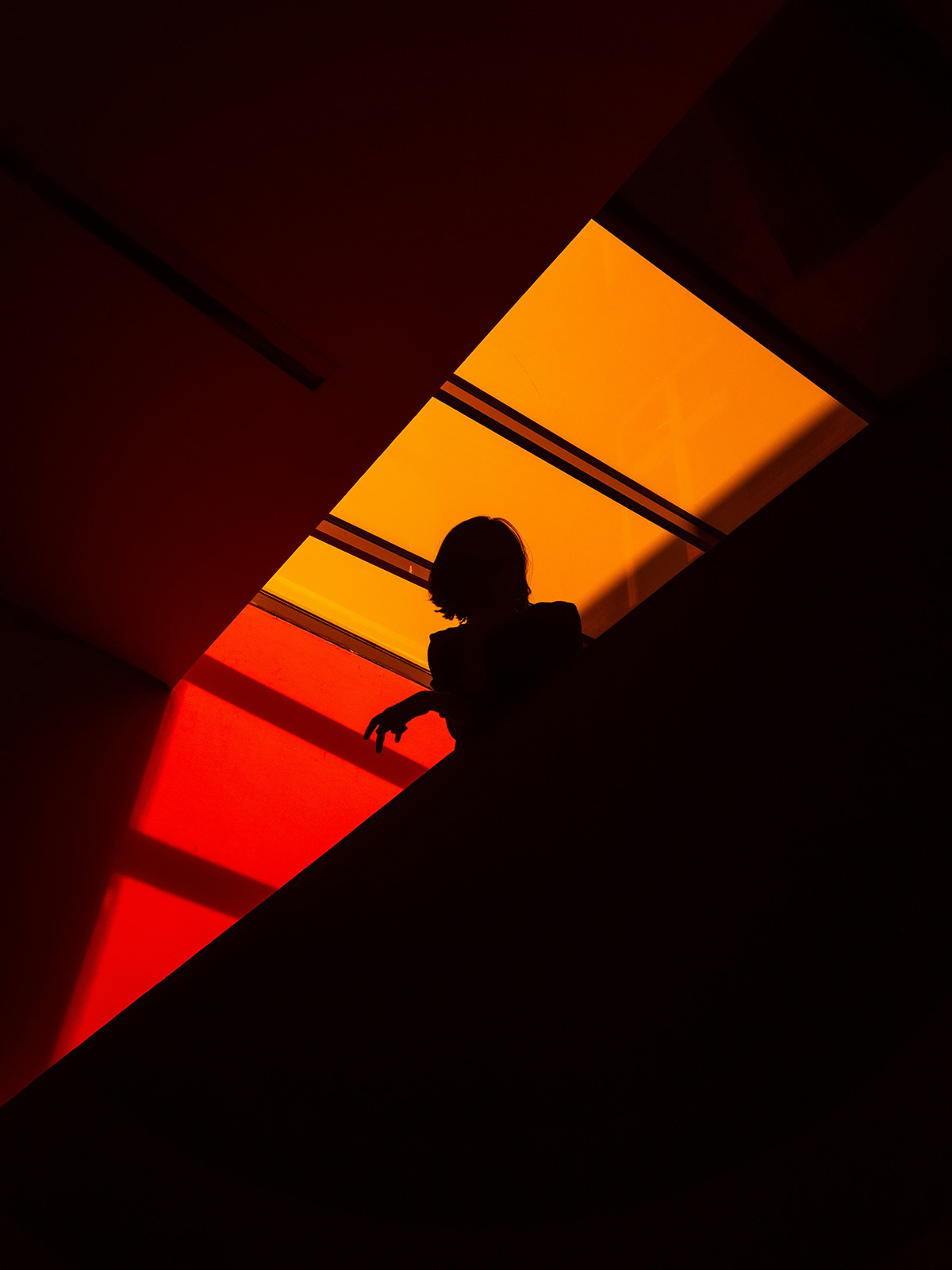 Silhouette of a person leaning on a railing, framed by dramatic red and orange lighting from a skylight above.